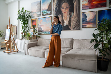 Obraz premium Thoughtful woman painter seeking inspiration stands with hand on chin. Contemplative girl in home art workshop among own paintings reflecting thinking immersing in dreams, fantasies, imagination.