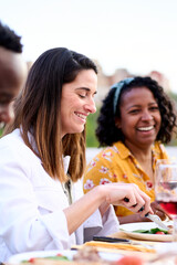 Vertical. Side view of smiling Caucasian woman holding fork and knife eating food sitting at table with friends unfocused. Happy people gathered to enjoy delicious meal in summer celebration outdoor