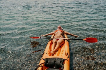 A woman is laying on a kayak in the water. The kayak is orange and has a paddle on it. The woman is...