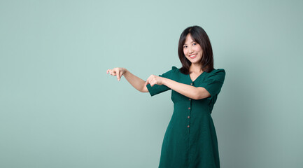 An Asian woman wearing a green dress is pointing to an empty space in a studio with a green background.