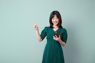 Asian woman green dress pointing finger to free copy space while using mobile phone isolated on pastel green background. online payment, promotion, deals concept.