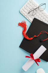 Overhead vertical view of a graduation cap, diploma with red ribbon, glasses, and a white keyboard...