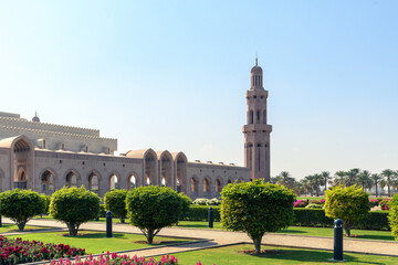 Obraz premium A serene view of the iconic Sultan Qaboos Grand Mosque in Muscat, Oman, showcasing its intricate Islamic architecture amidst lush green gardens under the clear blue sky.