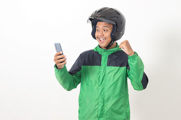 Portrait of Asian online taxi driver wearing green jacket and helmet using mobile phone while raising his fist and celebrating success. Isolated image on white background