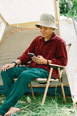 A real transgender Asian lesbian using Surf the web and social media on smartphone smiling in relaxation while sitting outdoors near a tent.