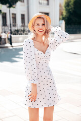 Young beautiful smiling blond woman in trendy summer white dress. Sexy carefree woman posing in street  at sunset. Positive model outdoors at sunny day. Cheerful and happy. In hat
