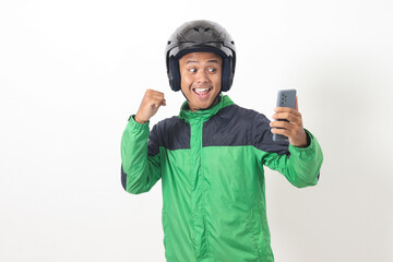 Portrait of Asian online taxi driver wearing green jacket and helmet using mobile phone while raising his fist and celebrating success. Isolated image on white background