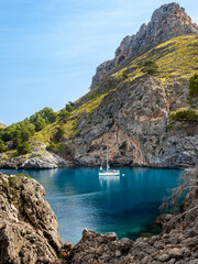 Morning allure of Cala de Sa Calobra bay, featuring an anchored sailboat in calm blue waters embraced by rocky cliffs with el Turmàs mountain in background, ideal for travel and tourism promotions.