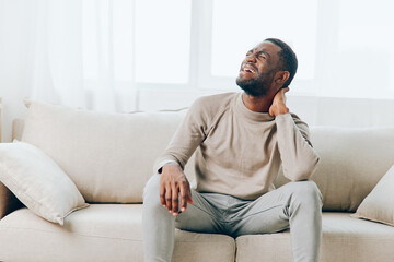 Tired African American man sitting on the couch at home, experiencing a painful headache His...