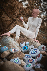 Young hairless girl with alopecia in white futuristic costume pensively examines stone with eye at...