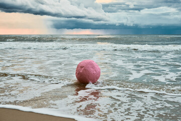 Large pink sphere lapped by waves against seashore against horizon and storm clouds, pink ball...
