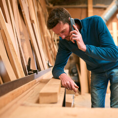 Carpenter Working In Woodwork Workshop Making Call On Mobile Phone Whilst Choosing Wood