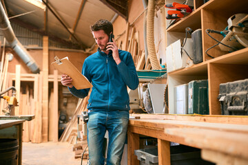 Carpenter Working In Woodwork Workshop Making Call On Mobile Phone Whilst Looking At Plan