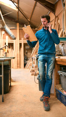 Carpenter Working In Woodwork Workshop Making Call On Mobile Phone Whilst Looking At Plan