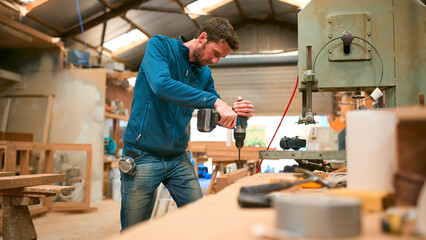 Carpenter At Workbench In Workshop Using Cordless Electric Drill