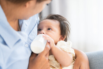 Asian woman feeding milk her cute mixed race little newborn 3 months old baby girl from bottle. Mother holding adorable small daughter in warm arms at home. Copy space