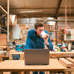 Carpenter Working In Woodwork Workshop Using Laptop With Cup Of Coffee