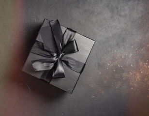 Black gift box with black bow on dark background with bokeh and sparkles. Top view, space for your text or design