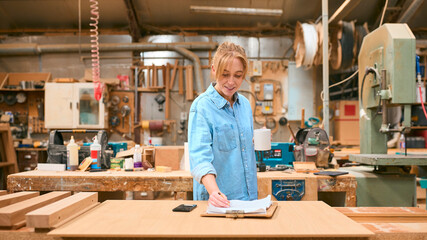 Female Carpenter Working In Woodwork Workshop With Cup Of Coffee Making Notes On Clipboard
