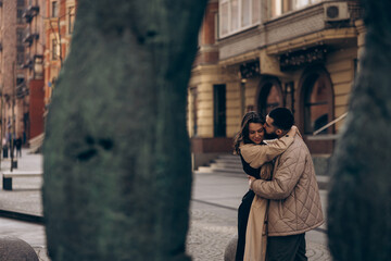 Happy young couple kissing and embracing against background of buildings and tree trunks.