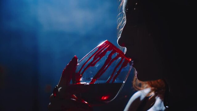 unrecognizable dark silhouette vampire woman licks drops of blood from lips beauty face sexy girl. hand holding glass red wine drinking bloody drink. art halloween holiday party. Gothic lady vamp 4k