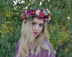 close up portrait of pretty blonde female model wearing a flower crown wreath and purple dress.  green nature  plants and trees in background