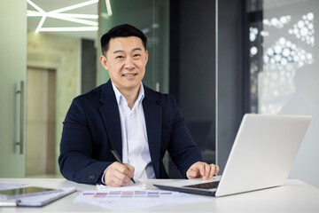 Asian mature man working on a laptop in a modern office setting, demonstrating productivity and professionalism.