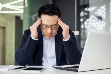 Asian mature man in a business suit experiencing stress and headache while working on a laptop at the office.