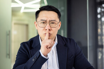 Asian mature man in business attire making a shushing gesture, emphasizing silence and...