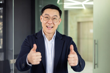 Smiling asian mature man wearing business attire giving thumbs up gesture in a modern office...