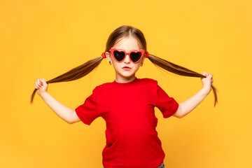The happy little girl in red t-shirt and red summer sunglasses lifts her ponytails of hair up, and...