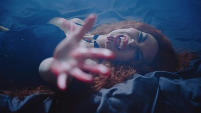 close-up fantasy vampire young woman lying on bed bedroom moon light night fog dark room shows sharp white teeth fangs lips blody beauty face waving hand in blood. sexy adult girl long red hair art 4k