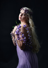 close up portrait of beautiful female model with long blonde hair, wearing purple fairy dress and fantasy flower crown. isolated against dark studio background, holding purple flowers