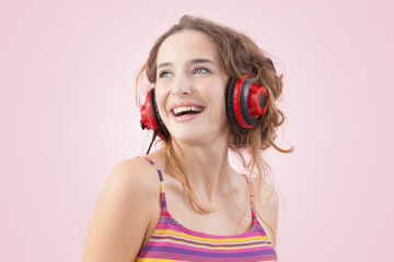Portrait of young joyful woman who is listening to music through red headphones, looks to the side with her blue eyes, while being isolated on a pink background