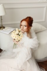 Radiant young woman with fiery red hair, dressed in white, striking a pose with a bouquet of white...