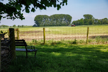 Tractor in a sunlit Dutch polder, with a shaded bench under trees.