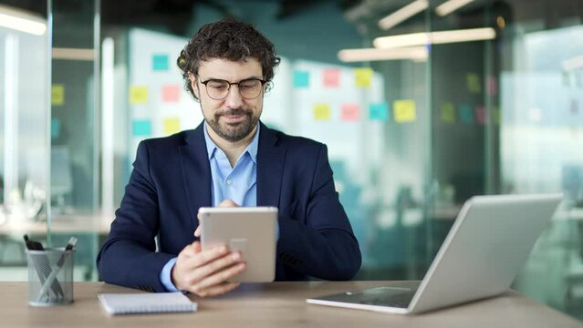 Confident businessman is using digital tablet while sitting at workplace in business office. Man worker works on application, typing a message, chatting with client, makes an order or browsing online