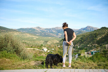Full size portrait of a woman in sportswear, walking her dog on leash in the nature. Multi ethnic...