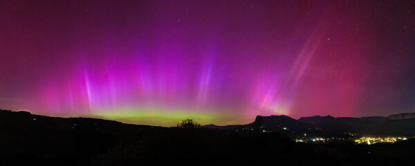 Panoramic night sky view of vivid aurora borealis above a mountainous landscape with glowing town...