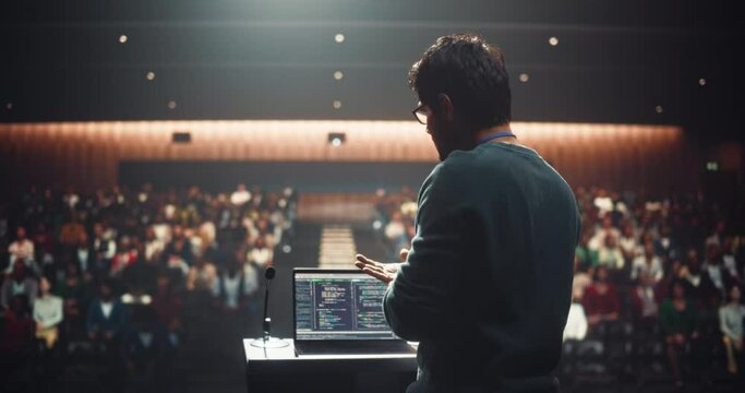 Young Empowering Thought Leader Standing Behind a Tribune, Showing an Inspiring Presentation About Software Innovations at Technology Conference. Indian Man Talking to an Audience in a Dark Venue
