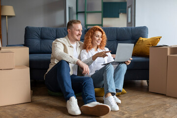 Couple using laptop computer sitting on floor among boxes after moving new home. Real estate, house ownership and relocation concept