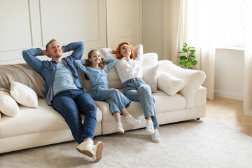 Relaxed parents and daughter resting on sofa with closed eyes and hands behind heads in living room...