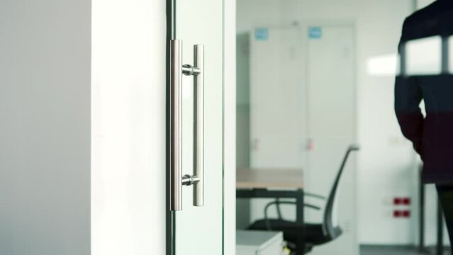Close up of a man's hand opening a glass office door holding a metal doorknob in a modern building. A man, businessman or employee in a formal suit opens the door and enters the workplace inside