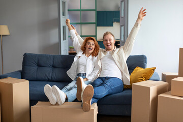 Excited husband and wife raise their hands up celebrating moving day, sitting on couch at new...