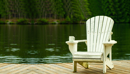 White Adirondack chair on wooden dock, with coffee mug on armrest. Serene water, lush trees in the background. Ample space for text.