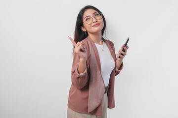 Cheerful Asian woman in eyeglasses holding smartphone and pointing to the copy space behind her,...