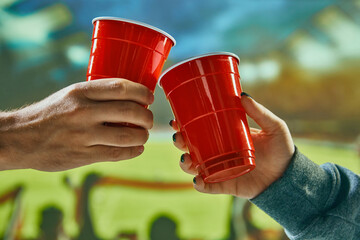 People clinking red mugs with beer at football stadium during match. Celebrating successful game....