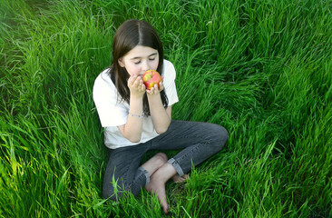 girl holding an apple in her hands and sitting on green grass, summer, happy childhood, health in nature, bare feet