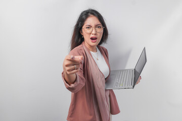 Enraged young Asian business woman holding a laptop pointing to the camera with angry expression, isolated by white background.
