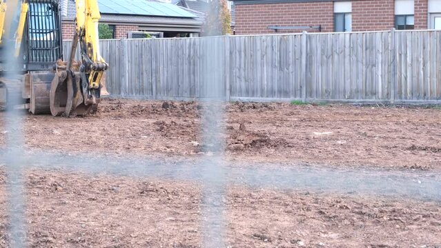 A vacant land covered with soil, sits a small excavator on a construction site for a future suburban home in a residential area, with fence and houses in the background neighborhood.